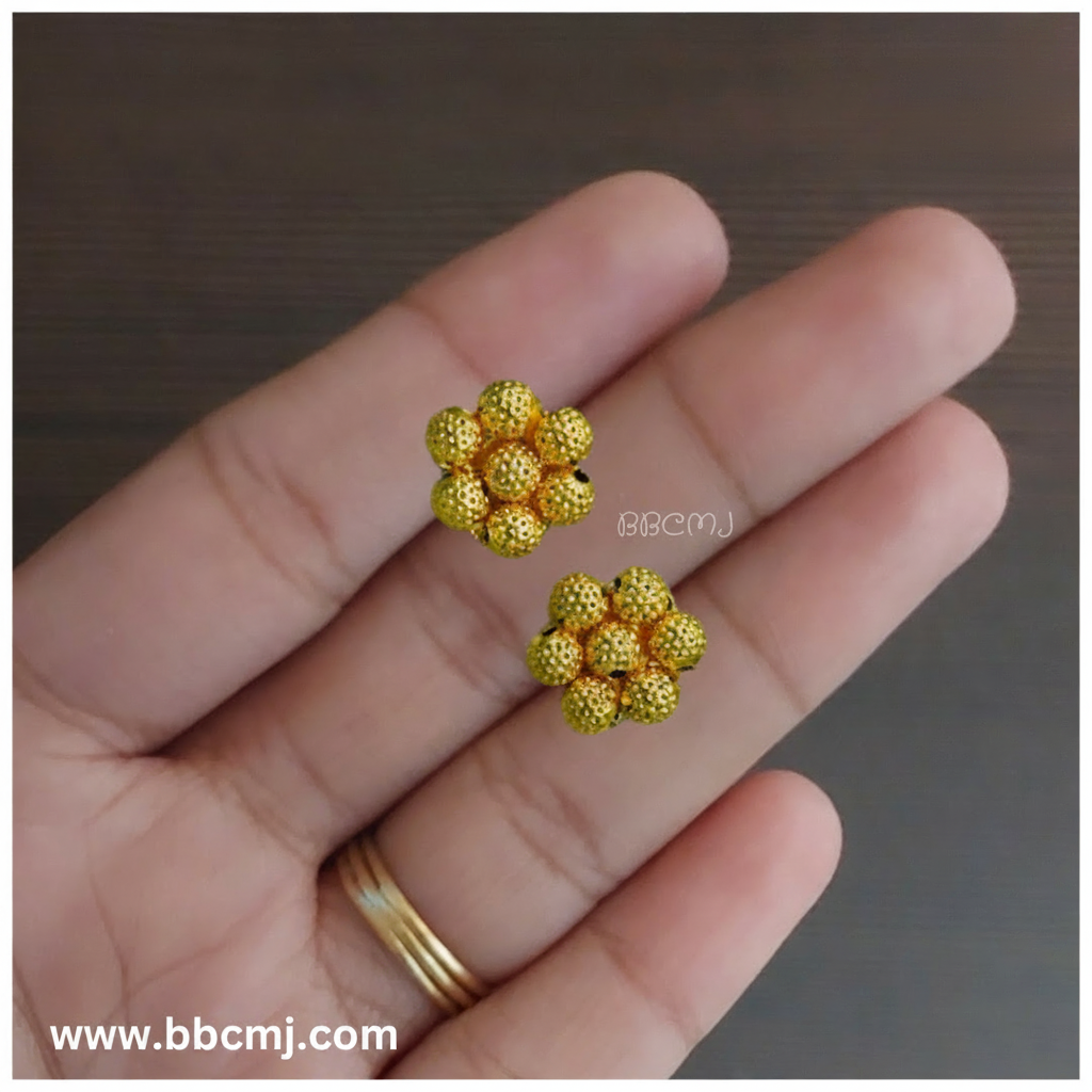 A pair of 1gm gold-coated earrings with a floral pattern, held in a person's hand, with the brand's website mentioned as 'bbcmj.com'.